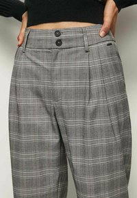 Grå rutig byxor med veckad front, två svarta knappar i midjan och en slät tygtextur med fina rutmönster.