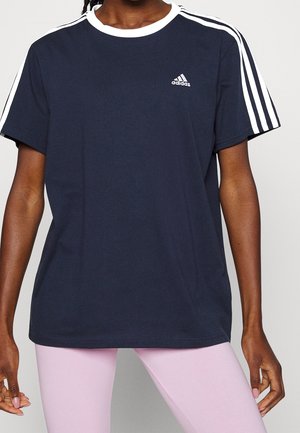 Personne portant un T-shirt Adidas bleu marine avec des bandes blanches sur les épaules et des leggings rose clair, debout devant un fond uni.
