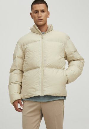 Beige pufferjakke med høj krave, store quiltede sektioner og frontlynlås. Blød, glat tekstur; stylet med beige bukser og en blå skjorte.
