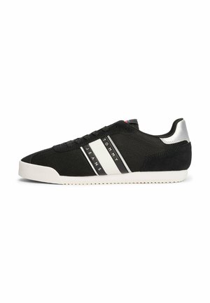 Zapatilla negra con material de malla y ante, rayas blancas y negras, parte superior texturizada, suela plana y acento plateado en el talón.