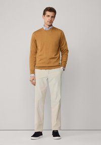 Musztardowy sweter, jasnoniebieski kołnierzyk koszuli, kremowe spodnie, czarne trampki. Sweter ma ściągacze przy dekolcie i rękawach oraz dopasowany krój.