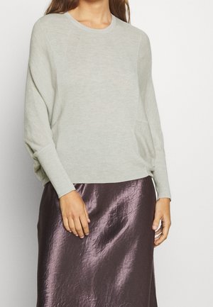 Femme portant un pull gris clair à manches longues et une jupe en satin violet foncé brillant sur un fond blanc uni.
