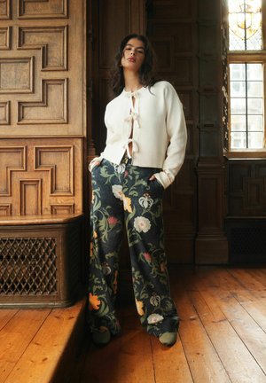 Eine Frau steht in einem holzvertäfelten Raum und trägt eine weiße Strickjacke mit Binderverschlüssen und weiten, floral gemusterten Hosen in Grün- und Orangetönen.