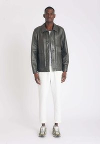 Olijfgroene leren jas met vier voorzakken, knoopsluiting en een losse pasvorm, gedragen over een lichtgrijs shirt en witte broek.