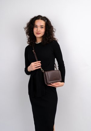 Brown leather crossbody bag with a smooth texture, a gold logo, and a strap featuring a knot detail, held by a person in a black dress.