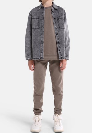 Barn iført en vaskeblød sort denimjakke over en brun sweatshirt og brune joggingbukser med hvide sneakers på hvid baggrund.