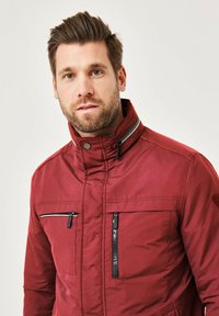 Red jacket with a high collar, featuring multiple zippered pockets and contrasting stitching. Material appears textured and durable.