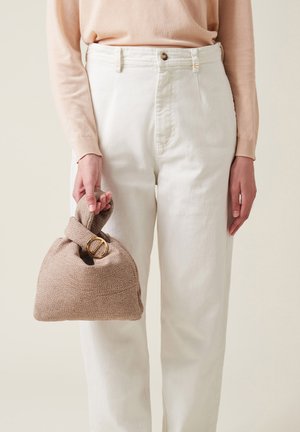 Sac à main en tissu beige texturé avec une poignée nouée et des accessoires en or, tenue par une personne portant un pantalon clair et un pull.