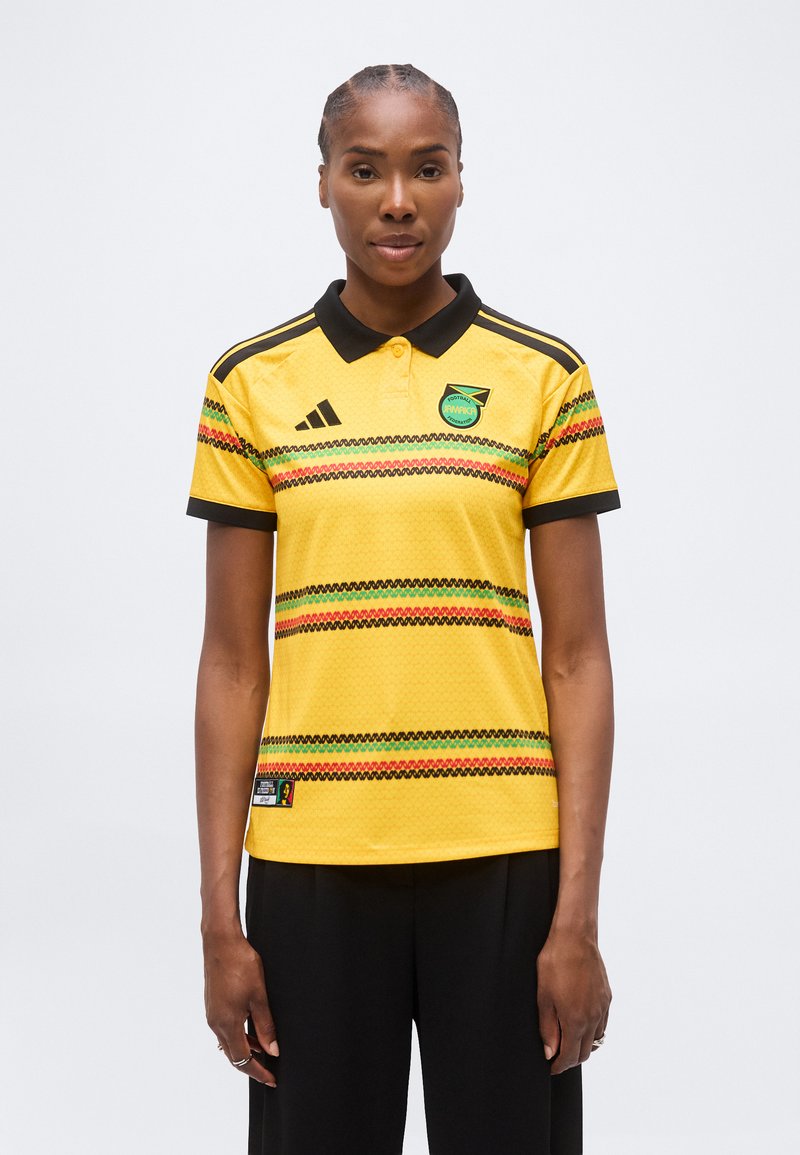 Γυναίκα που φοράει κίτρινη ποδοσφαιρική φανέλα της Τζαμάικας με μαύρο γιακά και πολύχρωμες ζιγκ ζαγκ ρίγες, στέκεται μπροστά σε απλό λευκό φόντο.