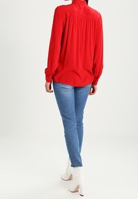 Blouse rouge à manches longues avec un dos froncé, associée à un jean skinny bleu et des bottines blanches, sur un fond uni.