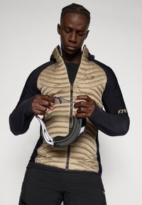 Veste isolée beige et noire avec fermeture éclair sur le devant, texture rembourrée et capuche ; lunettes de ski tenues avec une sangle blanche et un cadre noir.