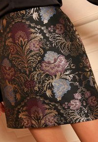 Fekete szövet szoknya bonyolult arany, burgundi, kék és rózsaszín virágmintás brokát mintával, egy személy alsó testén bemutatva.