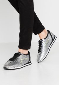 Sneaker metallici argento con una texture a forma di scaglie di pesce. Lacci e dettagli neri, con una striscia gialla brillante sul lato.