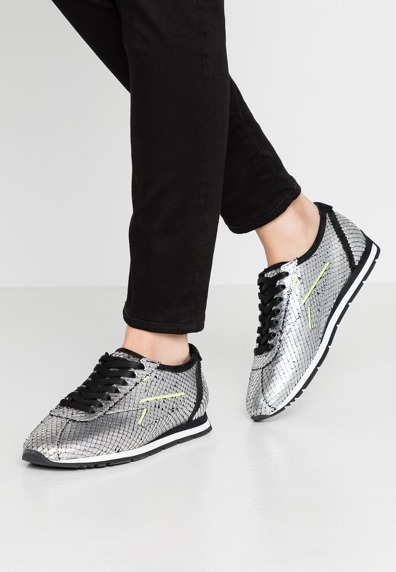 Sneaker metallici argento con una texture a forma di scaglie di pesce. Lacci e dettagli neri, con una striscia gialla brillante sul lato.