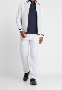 Vit lättviktsjacka med dragkedja över en marinblå skjorta, parat med vita byxor i rutigt mönster och svarta sportskor.