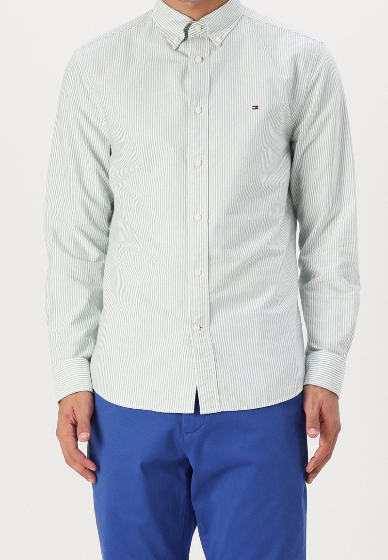 Homme portant une chemise blanche et gris clair à fines rayures avec un pantalon bleu, debout les bras détendus le long du corps.