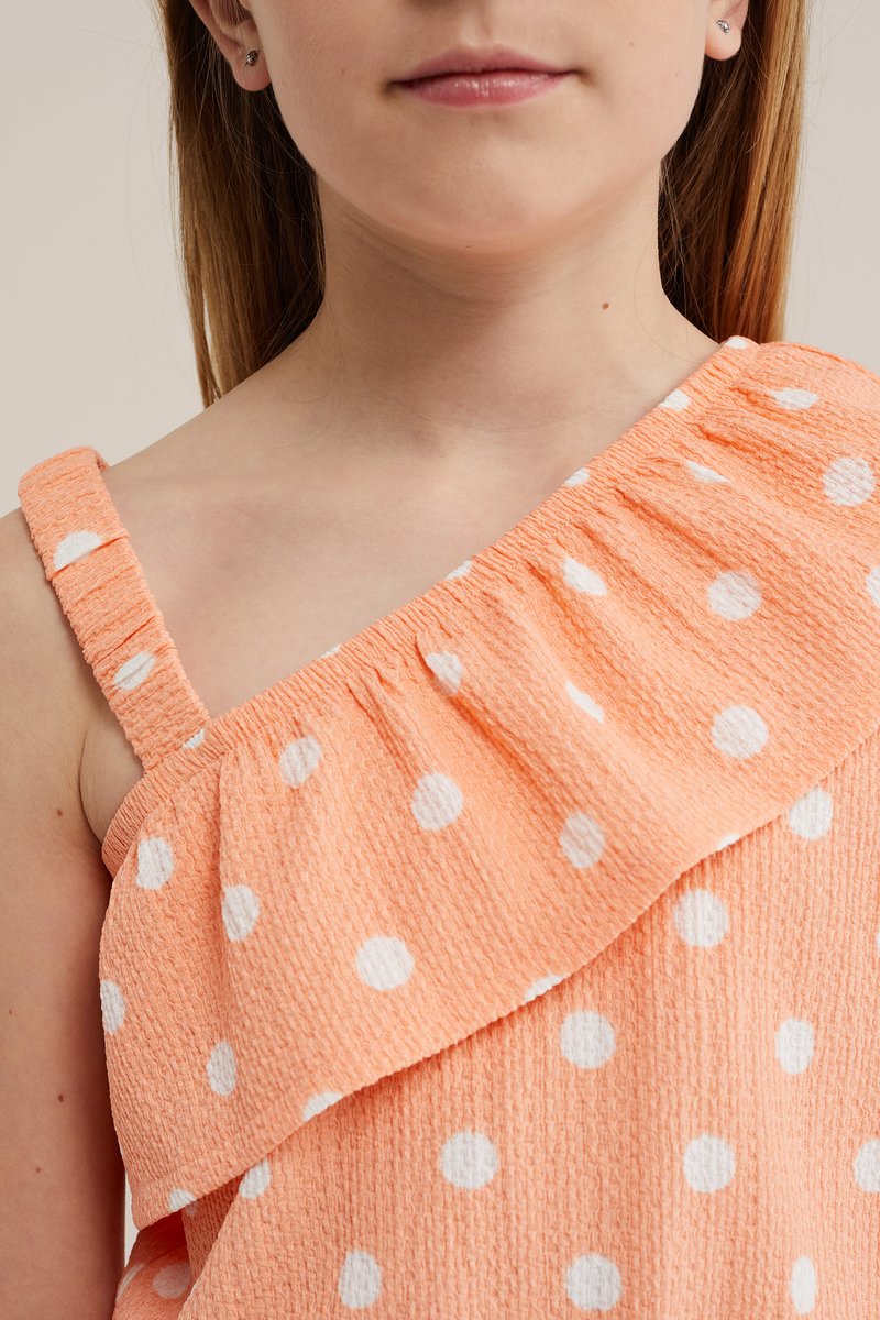Enfant portant une robe pêche asymétrique à pois blancs avec un détail froncé.