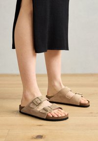 Sandales en daim beige avec deux brides réglables, semelle en liège et semelles en caoutchouc, mises en valeur sur des pieds avec un fond en bois.