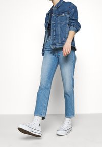 Spijkerjack in diepblauw met borduurdetails, gecombineerd met lichtblauwe jeans met rechte pijpen en witte hoge sneakers.