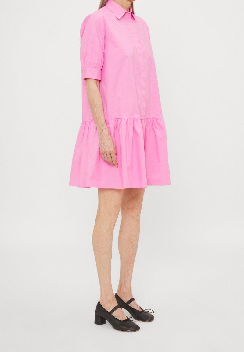 Femme portant une robe chemise rose vif ample avec des manches bouffantes jusqu'au coude et des chaussures Mary Jane noires à talon bas.