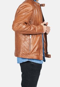Braune Lederjacke mit einem gesteppten Design, ausgestattet mit einem Stehkragen, Reißverschlusstaschen und einer glatten Textur. Trägt man über einem blauen Hemd.