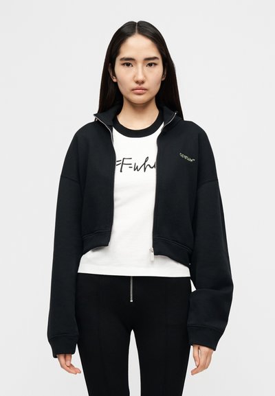 Μαύρο cropped φούτερ με φερμουάρ, χοντρά μανσέτα και ψηλό γιακά, σε συνδυασμό με λευκό t-shirt με μαύρο κείμενο. Ομαλή υφή υφάσματος.