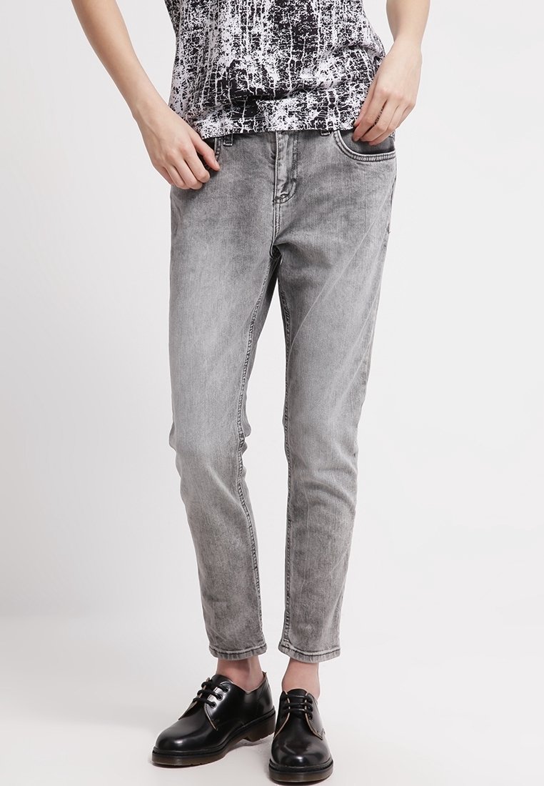 Jean de mezclilla gris con un corte entallado, textura ligeramente desgastada y costuras visibles. Combinado con zapatos de cuero negro y una blusa de patrón en blanco y negro.