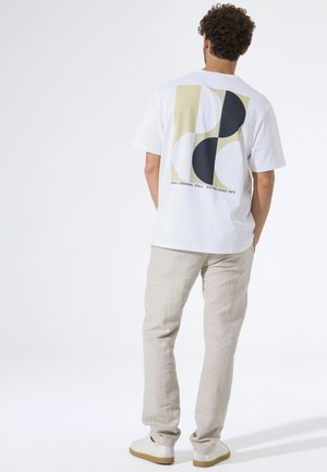White cotton t-shirt with abstract design in mustard, black, and white on back, paired with light beige linen pants and white sneakers.