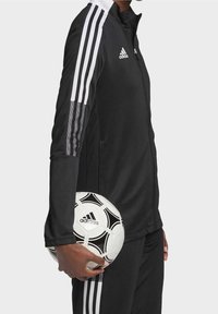 Person in black Adidas tracksuit holding a black and white Adidas soccer ball at their side, against a plain background.