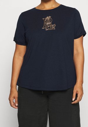 Kvinde iført en navyblå kortærmet top med opbukket kant og et gyldent broderet monogram på brystet, kombineret med sorte bukser.