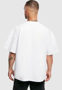 Fehér pamut, oversize póló rövid ujjakkal, egyenes hemeléssel és minimális varrási részletekkel. Látható tetoválások a bal karon.