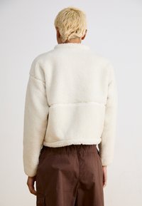 Weiße, flauschige Fleecejacke mit cropped Design und geripptem Saum. Verfügt über lange Ärmel und einen Kragen, kombiniert mit braunen Hosen.