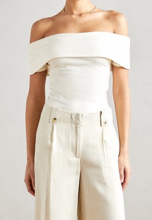 Kvinde iført en hvid off-shoulder top, stoppet ind i beige bukser med høj talje og vide ben med knapdetaljer ved taljen.