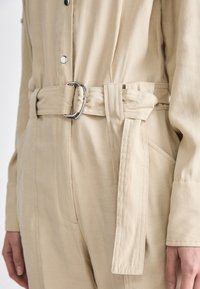 Combinaison beige avec une taille ceinturée mettant en valeur une boucle en métal argenté. Le tissu est lisse avec des coutures visibles et des poches latérales.