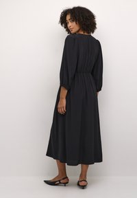 Robe midi noire à manches longues, taille froncée et tissu fluide. Texture lisse avec de subtils rayures verticales, associée à des escarpins noirs à kitten heels.