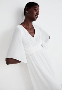 Λευκό φόρεμα με V λαιμόκοψη, κοντά φουσκωτά μανίκια, και ελαστική μέση. Φτιαγμένο από ελαφρύ ύφασμα με ροή στην φούστα.