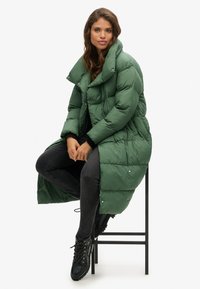 Lunga giacca imbottita verde scuro, con colletto alto, motivo trapuntato e chiusura a bottoni a pressione. Abbinata a jeans scuri e stivali neri.