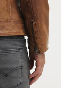 Brun læderjakke med en tekstureret finish, der har lynlåslukning på manchetterne, kombineret med grå denimjeans.