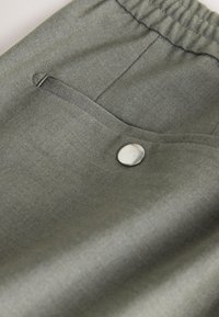 Gros plan sur un tissu gris avec une ceinture élastique, une poche passepoilée et un détail d’un seul bouton métallique brillant.