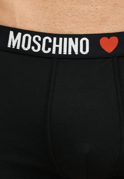 Μαύρη ζώνη εσωρούχου με λευκή επιγραφή "MOSCHINO" και κόκκινο σύμβολο καρδιάς πάνω σε γυμνό δέρμα.