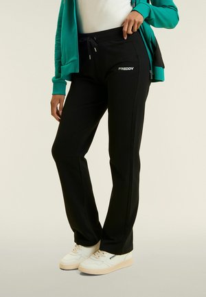 Persona che indossa pantaloni della tuta neri del marchio Freddy, scarpe da ginnastica bianche e una giacca verde con cerniera, in piedi con una mano sul fianco.