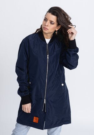 Granatowa kurtka bombowca z zamkiem błyskawicznym, ribbowymi mankietami, przednimi kieszeniami oraz pomarańczowym detalem na dole. Gładka, błyszcząca tekstura.