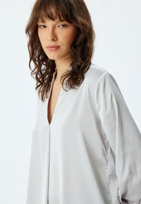 Zijdezachte, lichtgrijze blouse met een V-hals, lange mouwen en een ontspannen pasvorm. Heeft subtiele plooidetails aan de voorkant.