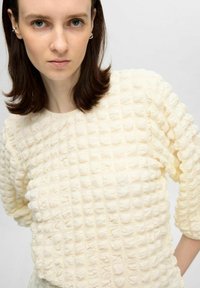 Krämfärgad topp med en texturerad, vadderad mönstring. Har en rund halsringning och ärmar i tre fjärdedelar. Tillverkad av bomullsblandning.