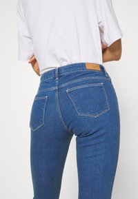 Osoba v modrých džínách Wrangler s vysokým pasem a bílou košilí s krátkým rukávem, zobrazená zezadu.