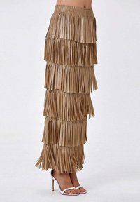 Jupe longue à franges couleur beige, fabriquée en matériau texturé. Présente de multiples détails de franges superposées. Associée à des sandales à talons hauts blanches.