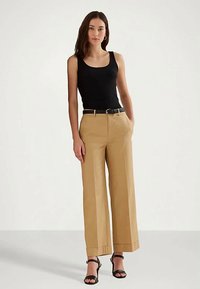 Γυναίκα που φοράει μια μαύρη αμάνικη μπλούζα, μπεζ παντελόνι με φαρδιά πόδια και μαύρη ζώνη, καθώς και μαύρα τακούνια, που στέκεται μπροστά σε λευκό φόντο.