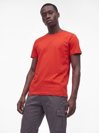 Mand iført en ensfarvet rød kortærmet t-shirt og grå cargobukser står foran en hvid baggrund.