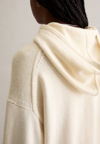 Felpa con cappuccio lavorata a maglia color crema, caratterizzata da una texture morbida, una vestibilità rilassata, un cappuccio drappeggiato e cuciture dettagliate lungo le spalle.