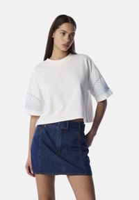 Witte cropped t-shirt met blauwe borduursels op de mouwen, gecombineerd met een donkerblauwe denim mini-jurk met een frontale knoopsluiting en riem.